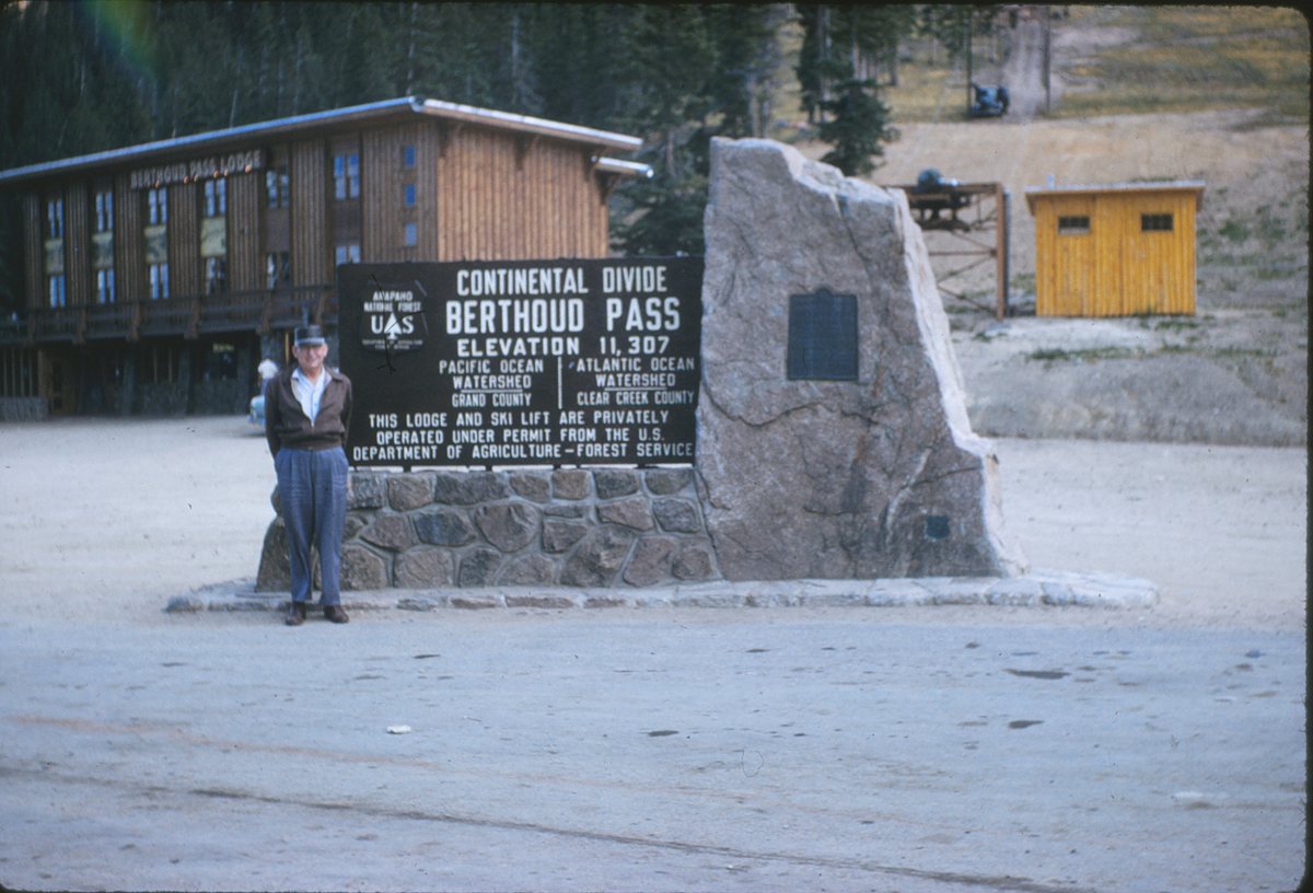Circa 1962 color photo at the Berthoud Pass summit: a man stands beside the Forest Service sign reading Continental Divide, Berthoud Pass, Elevation 11,307, with the Berthoud Pass Lodge and the ski lift terminal behind it