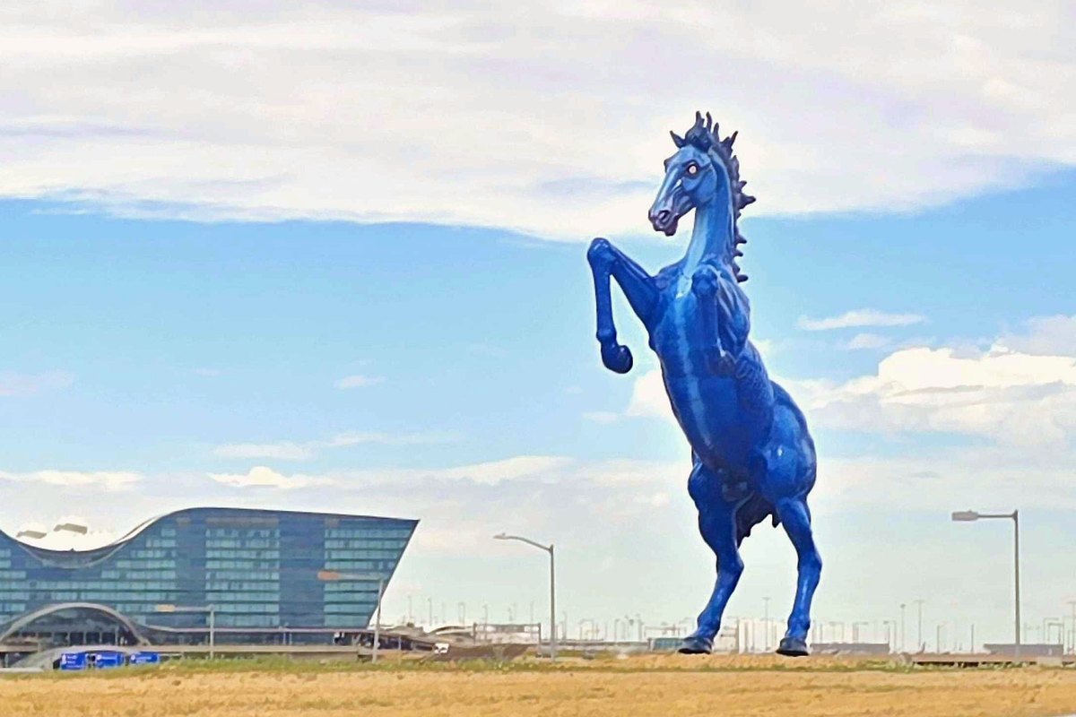 Mustang, the 32-foot blue horse sculpture with glowing red eyes, rearing beside Pena Boulevard with the Westin hotel and Denver International Airport control tower behind it